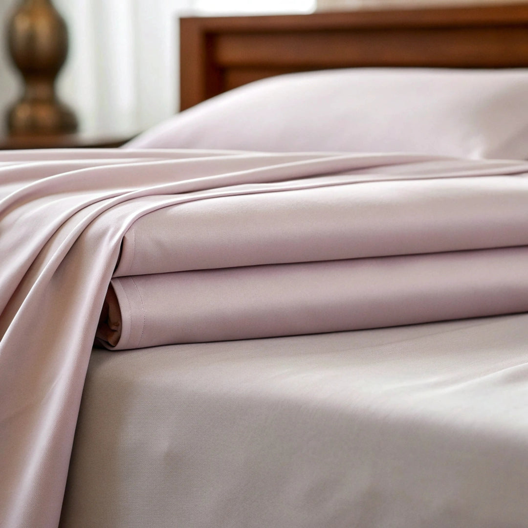 Close-up of pale light mauve rose bamboo sheets softly draped, highlighting their natural breathability and moisture-wicking properties. The blurred walnut bed frame in the background adds warmth, emphasizing a clean, healthy, and luxurious sleep environment.