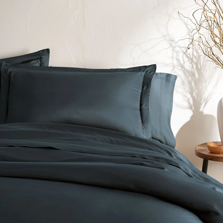 angled top-down view of slate bamboo sateen pillowcases and duvet on king bed, styled with neutral wall, ceramic vase, and wooden bedside table