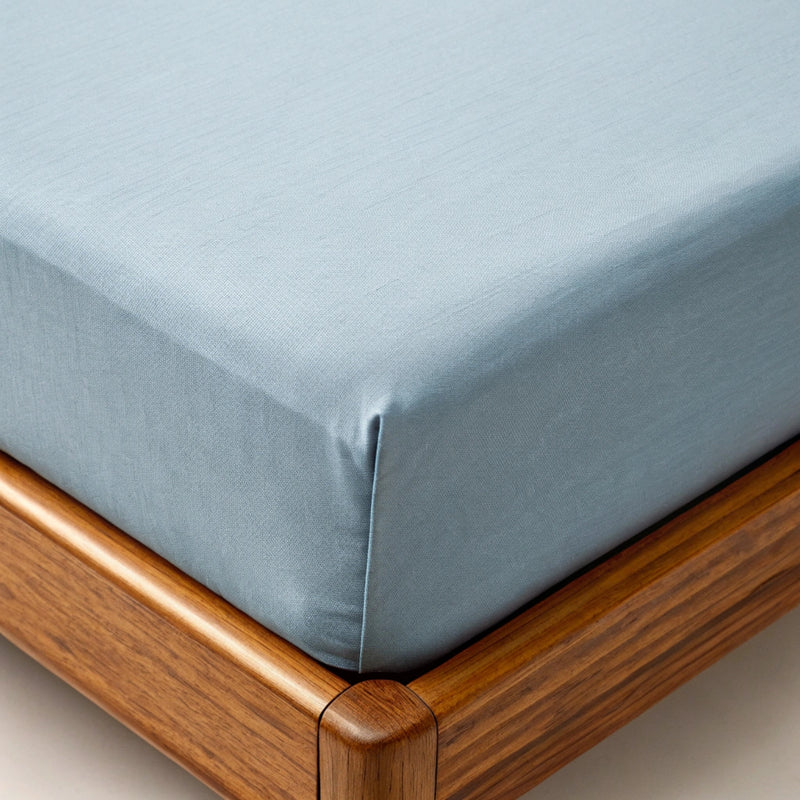 Close-up of dusty blue bamboo fitted sheet being pulled over mattress corner, showing soft twill weave, elastic fit, and walnut bed frame in background