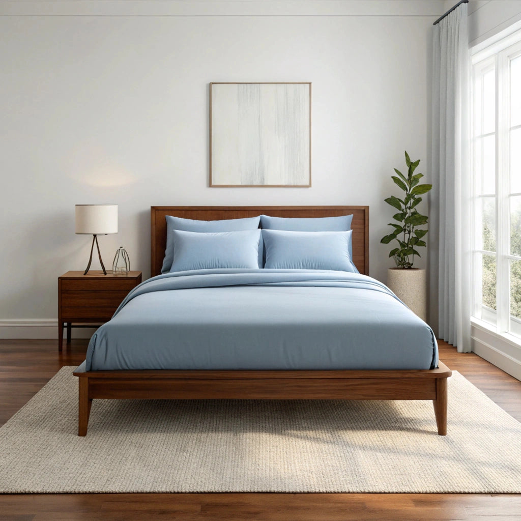 Wide-angle view of bedroom with dusty blue bamboo sheets on walnut bed frame, featuring matching nightstand, ceramic lamp, glass vase with greenery, soft rug, and minimal wall art