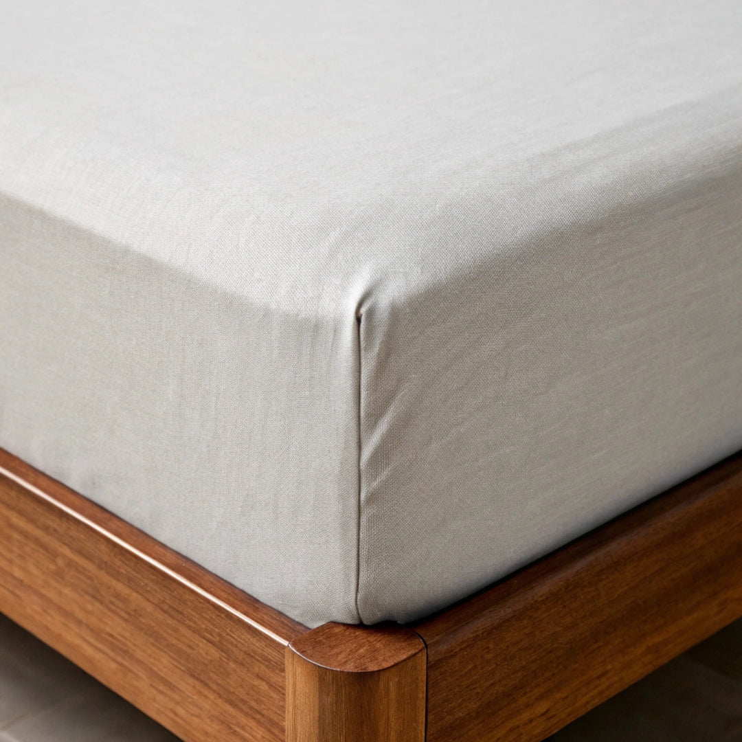 Close-up of feather gray  bamboo fitted sheet being pulled over mattress corner, showing soft twill weave, elastic fit, and walnut bed frame in background