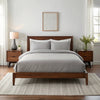 Wide-angle view of bedroom with feather gray bamboo sheets on walnut bed frame, featuring matching nightstand, ceramic lamp, glass vase with greenery, soft rug, and minimal wall art.