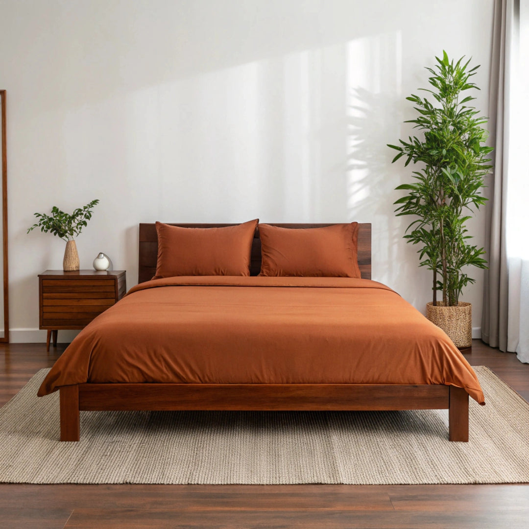Wide-angle view of bedroom with rust bamboo sheets on walnut bed frame, featuring matching nightstand, ceramic lamp, glass vase with greenery, soft rug, and minimal wall art.
