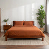 Wide-angle view of bedroom with rust bamboo sheets on walnut bed frame, featuring matching nightstand, ceramic lamp, glass vase with greenery, soft rug, and minimal wall art.