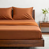 Side angle shot from pillow-end showing rust bamboo pillowcases and sheet set with decorative plant and soft lighting.