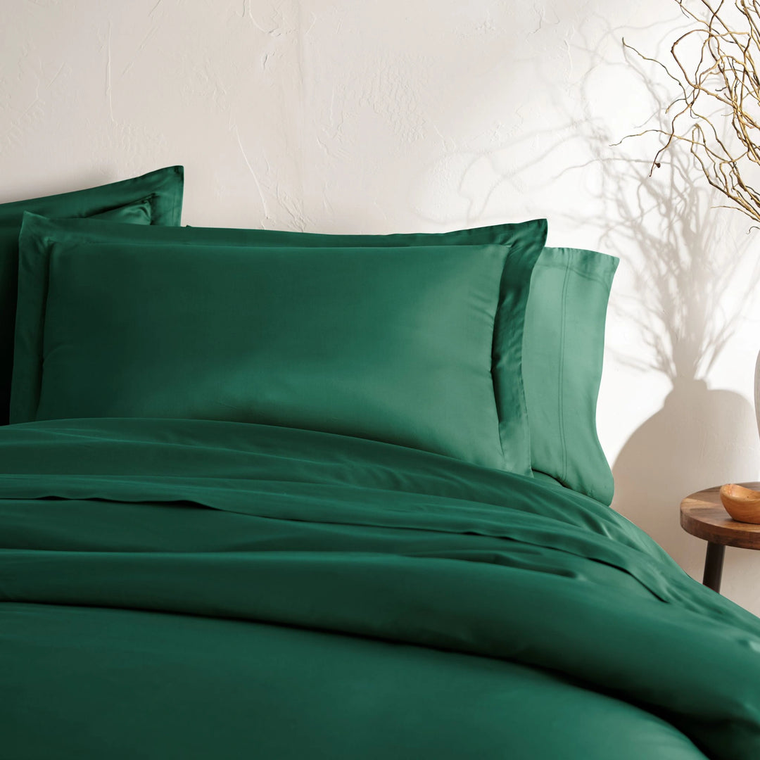 angled top-down view of emerald green bamboo sateen pillowcases and duvet on king bed, styled with neutral wall, ceramic vase, and wooden bedside table