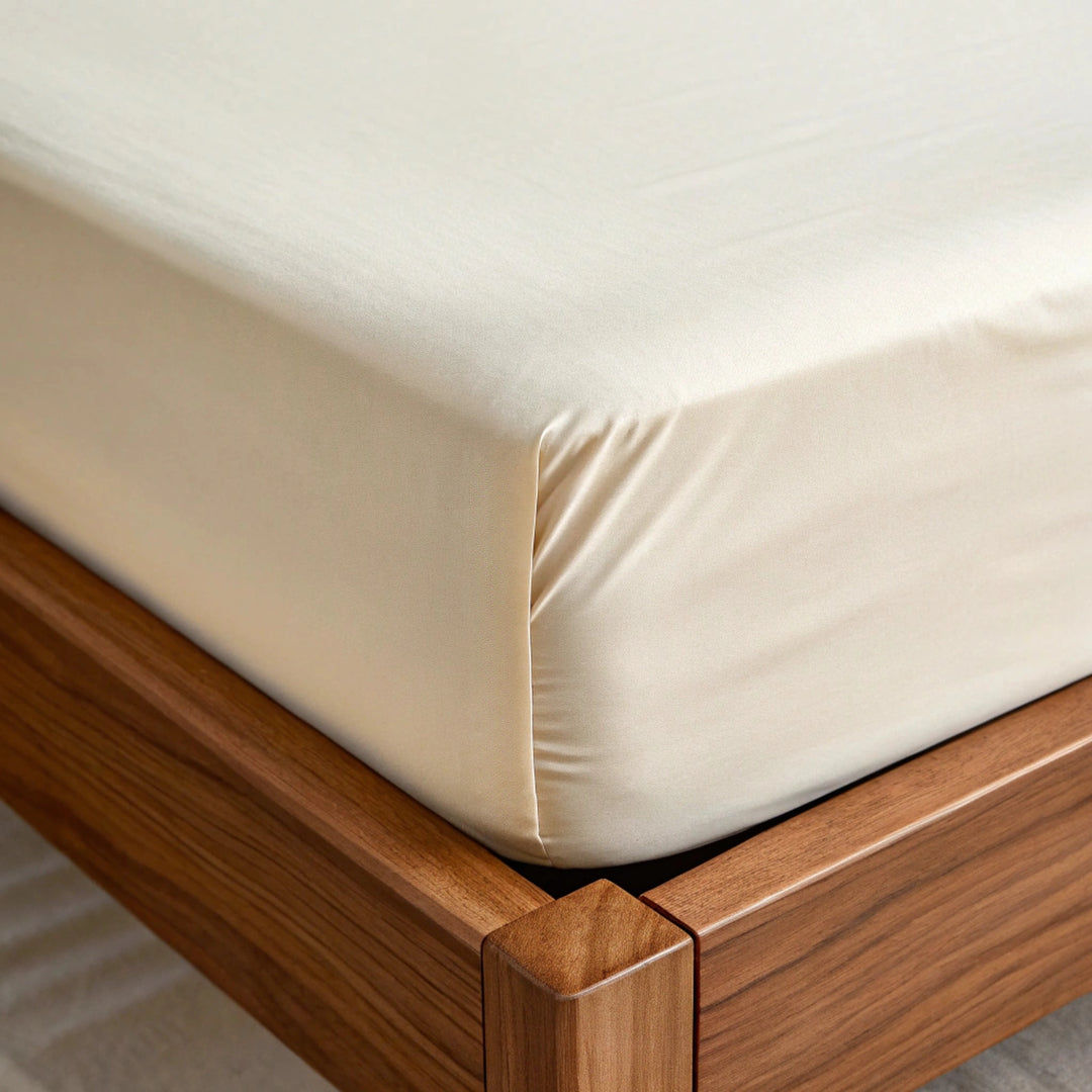 Close-up of a fitted ivory bamboo sateen sheet with twill weave being gently pulled over a mattress corner, highlighting the soft fabric and secure elastic fit. The warm walnut bed frame contrasts with the fabric, while natural light emphasizes the sheet’s stretch and clean, seamless design focused on everyday functionality.