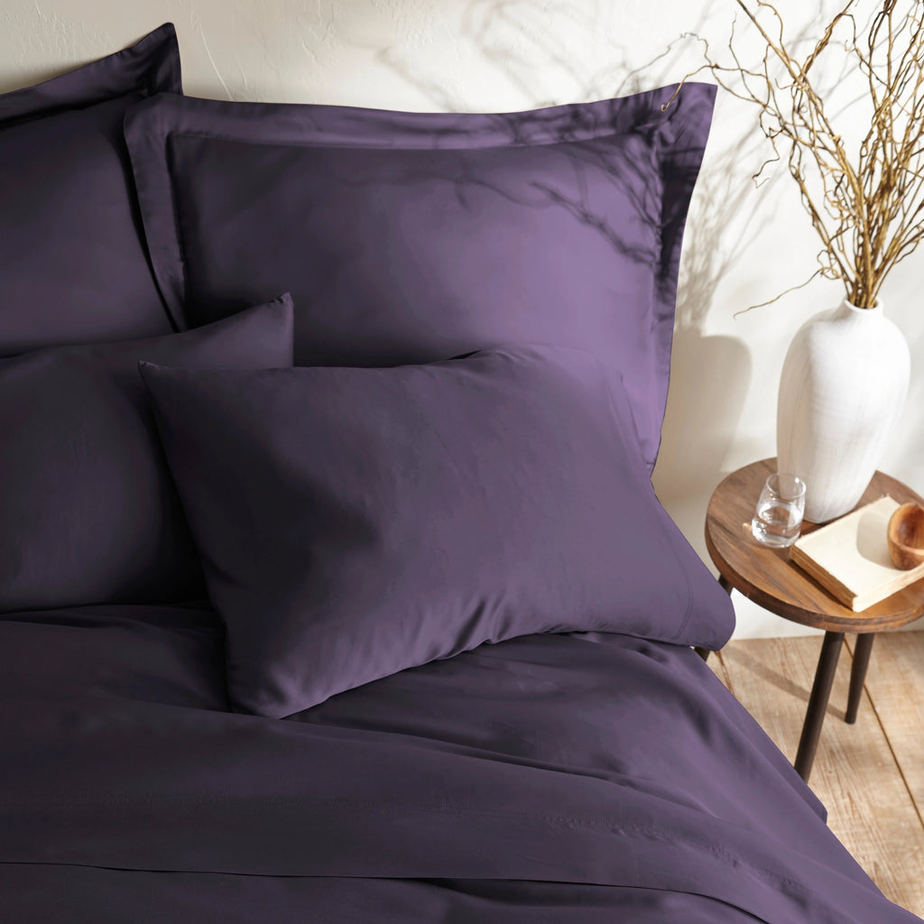 close-up of royal purple bamboo sateen pillowcases and duvet cover on king bed, styled against a textured cream wall with natural decor
