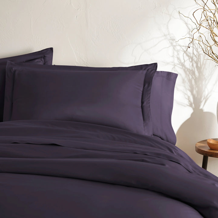 angled top-down view of royal purple bamboo sateen pillowcases and duvet on king bed, styled with neutral wall, ceramic vase, and wooden bedside table
