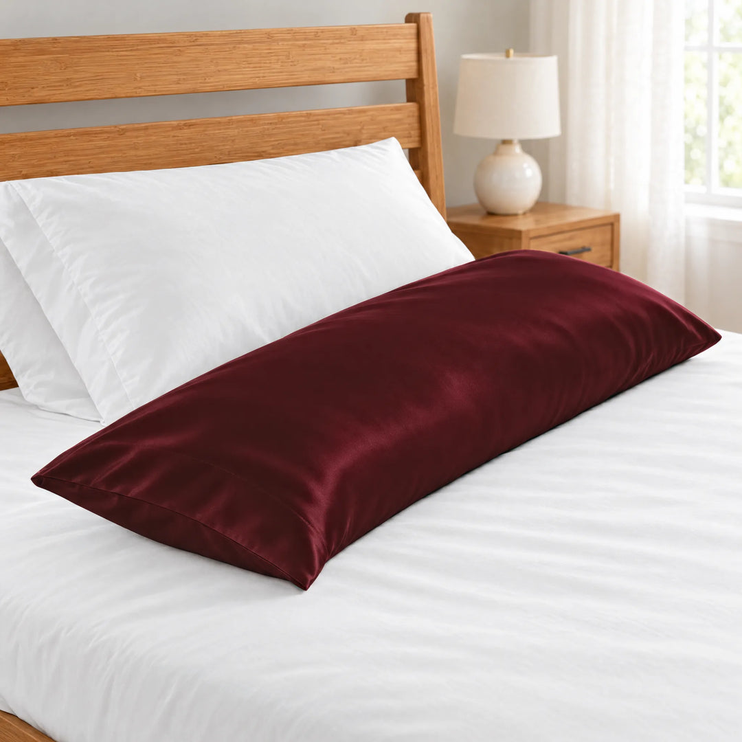 Ruby Red bamboo viscose sateen body pillowcase close-up of the stitched hem beside a white pillow on a made bed, honey bamboo headboard and lamp in soft focus