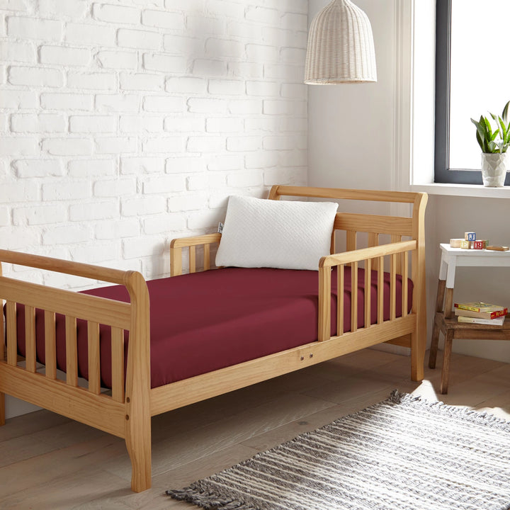 ruby red bamboo toddler fitted sheet from the Signature Collection, styled in a wooden toddler bed with ruby red pillow and modern nursery decor