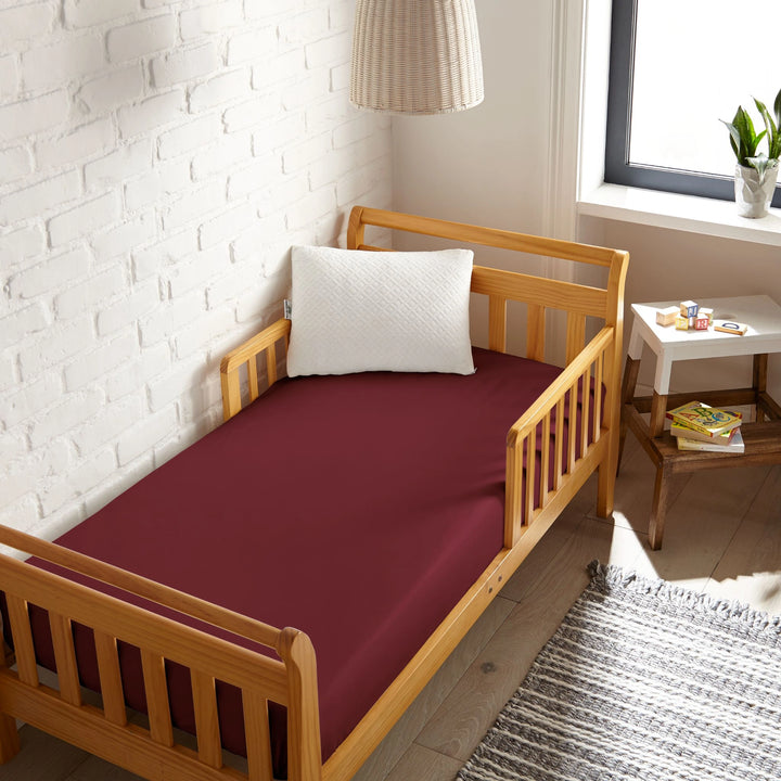 ruby red bamboo toddler fitted sheet from the Signature Collection, viewed from above on a light wood frame toddler bed in a bright modern nursery