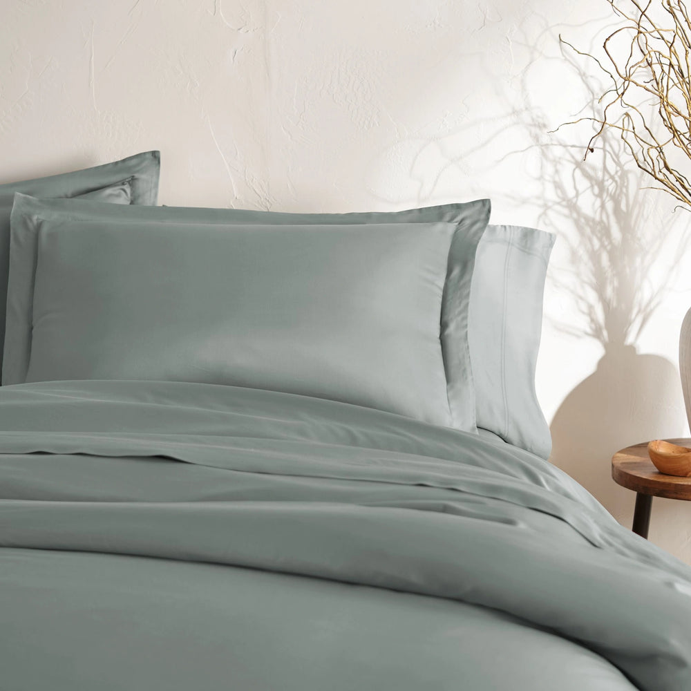 angled top-down view of silver bamboo sateen pillowcases and duvet on king bed, styled with neutral wall, ceramic vase, and wooden bedside table