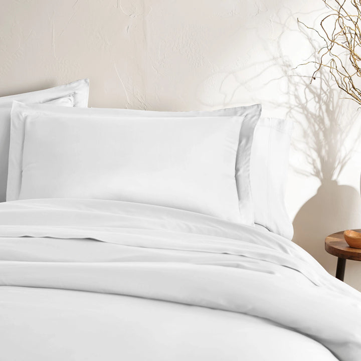 angled top-down view of white bamboo sateen pillowcases and duvet on king bed, styled with neutral wall, ceramic vase, and wooden bedside table