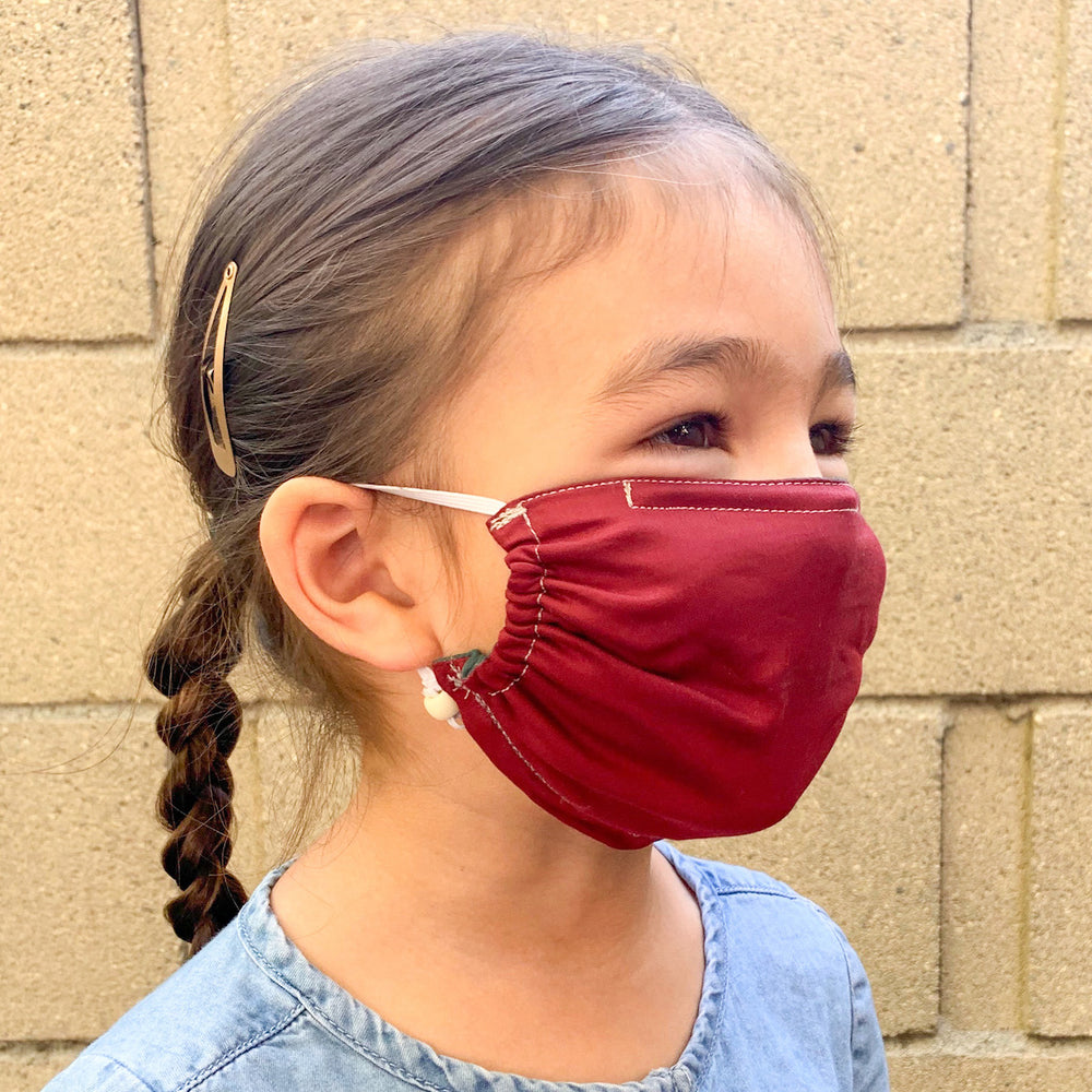 Young girl wearing a red face mask with a braided hairstyle, smiling outdoors with a beige wall in the background.