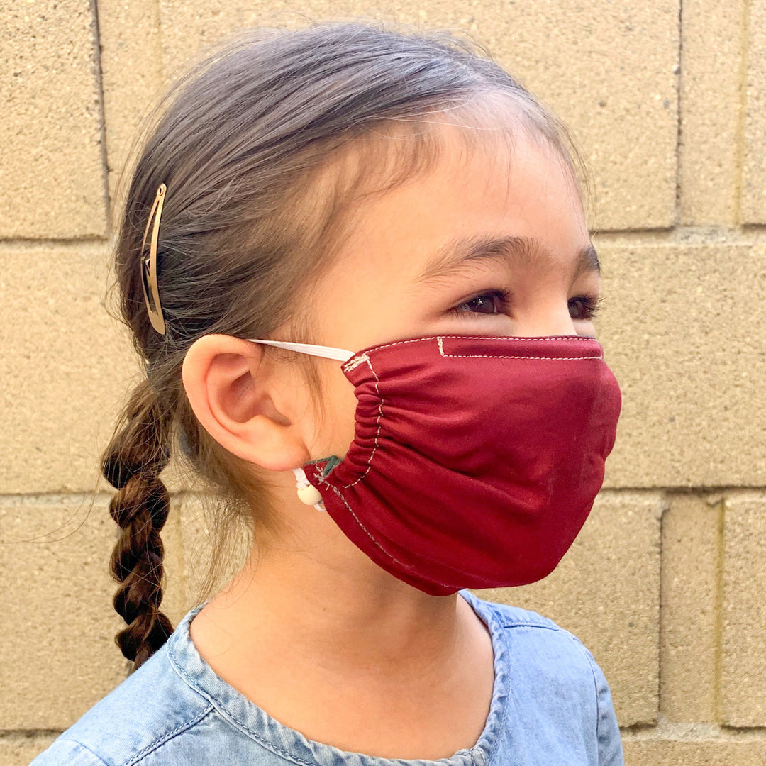 Young girl wearing a red face mask with a braided hairstyle, smiling outdoors with a beige wall in the background.